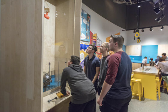 Photo of youths at a science exhibit where a hammer smashes safety helmets