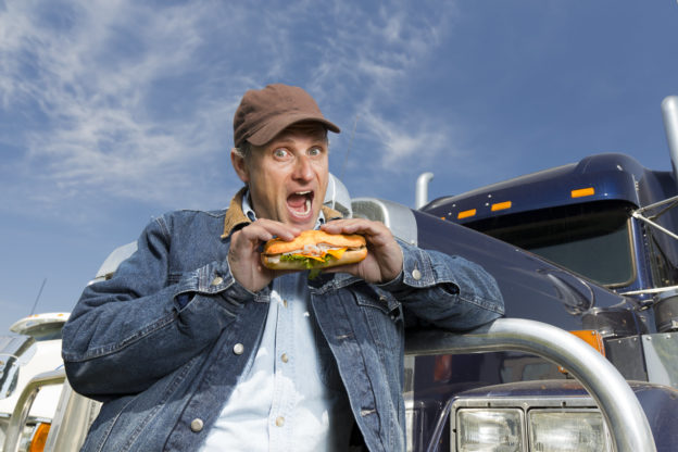Photo of truck driver eating loaded sub sandwich in front of truck.