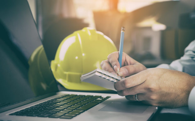 Photo of laptop, notebook, hands with pencil, and hard hat