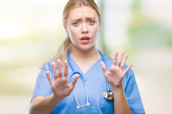 Photo of a blonde health care worker in blue scrubs looking afraid and holding her hands up palm outward