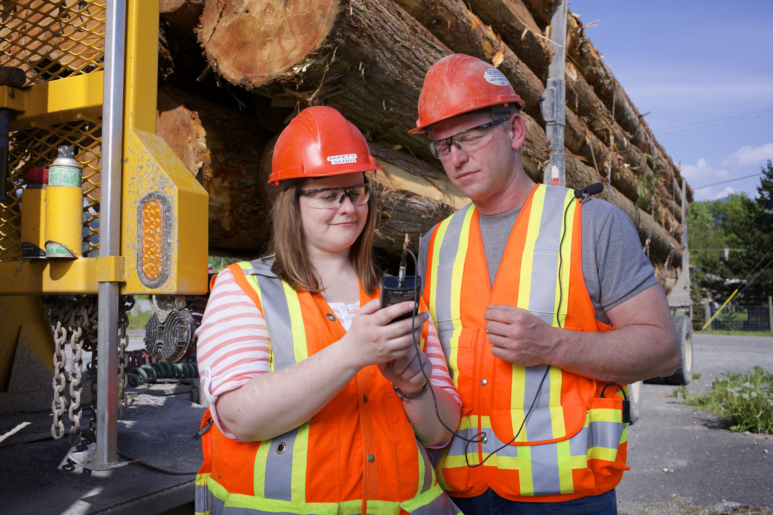 Photo of woman and man in safety vests and hard hats with loaded log transporter behind them