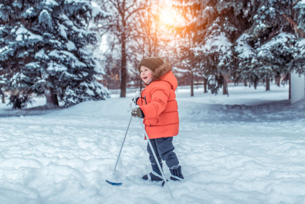 Photo of young child in winter gear, using skis on flat ground covered with snow, and nearby trees covered in snow