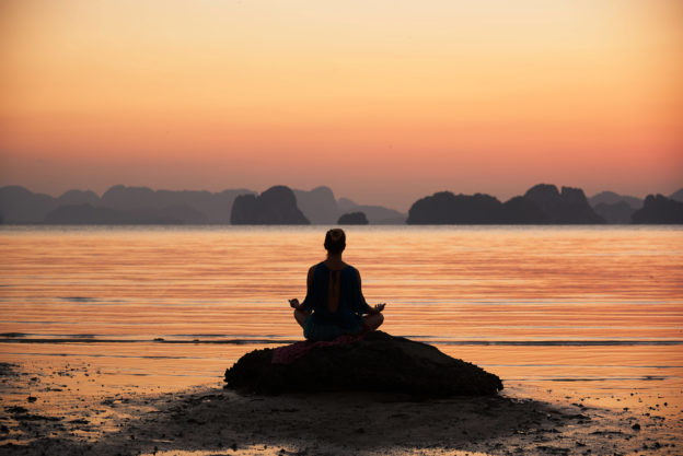 Photo of a figure meditating on a rock by the ocean during an orange sunrise