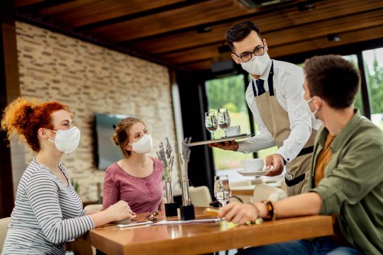 Photo of waiter serving coffee to group of friends while everyone one is wearing protective face mask due to COVID-19 epidemic.