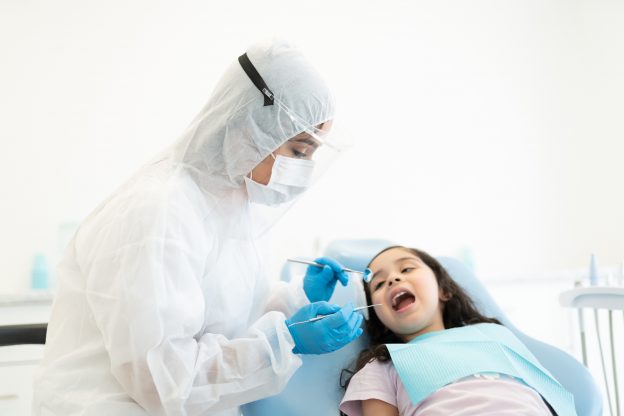 Photo of dentist in protective workwear checking teeth of girl with dental mirror and carver during COVID-19 pandemic