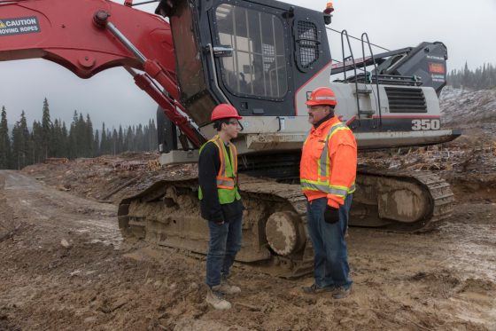 Photo of young worker interacting with older worker in front of a feller buncher