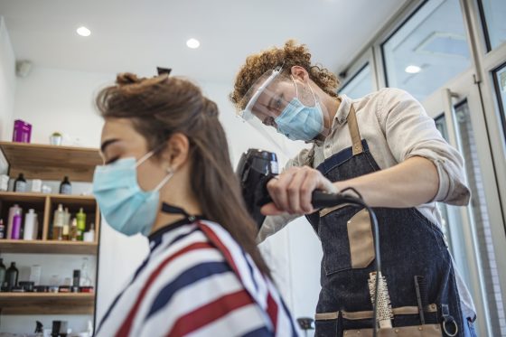 Photo of a hair stylist in a hair salon, wearing a mask and face shield, drying a customer's hair who is also wearing a mask.