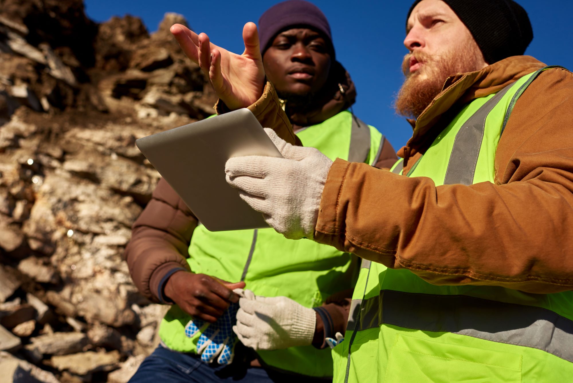Photo of two two industrial workers wearing reflective jackets, using a tablet and inspecting the land outdoors