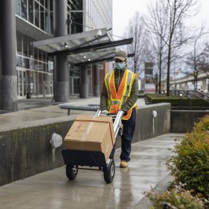 Photo of courier pushing a box on a dolly, down a path, while wearing a hi-vis vest and protective face mask.