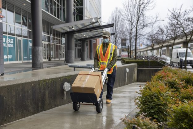 Photo of courier pushing a box on a dolly, down a path, while wearing a hi-vis vest and protective face mask.
