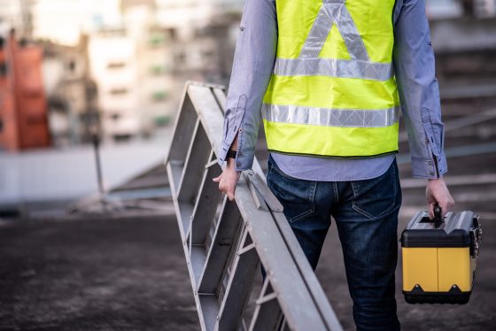 Photo of worker wearing a yellow vest, carrying aluminium stepladder and tool box at construction site