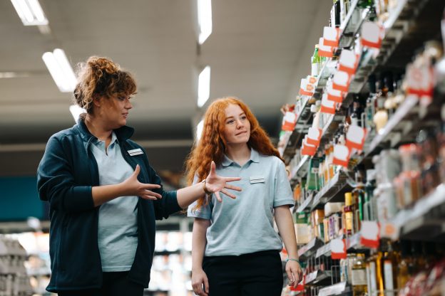 Photo of a supervisor training a young worker as they walk down the aisle of a grocery store.