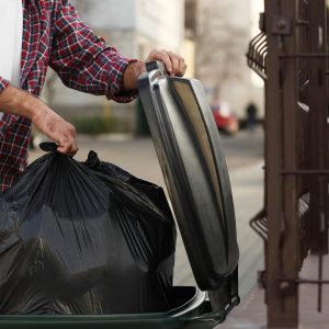 Photo of Man putting garbage bag into recycling bin outdoors, closeup