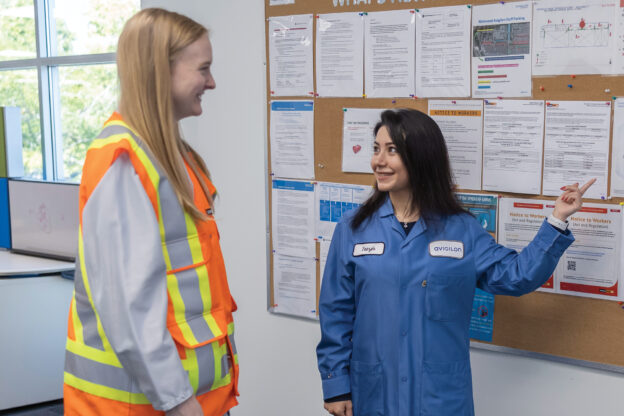 Photo of two women standing in front of a health and safety bulletin board having a conversation.