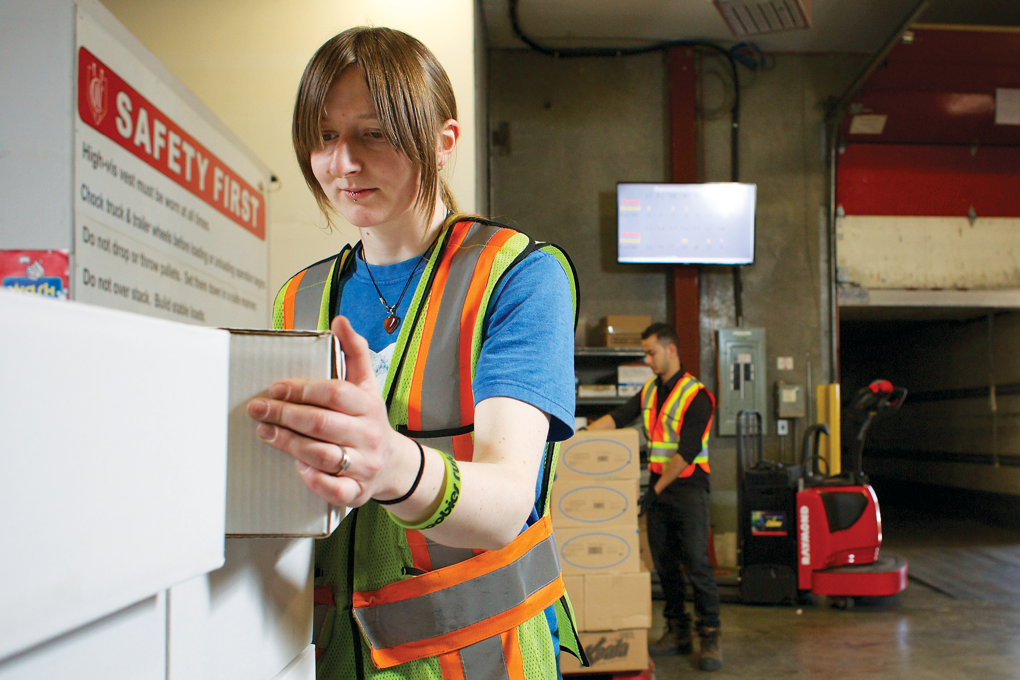 Photo of a female worker in a warehouse wearing a high vis vest picking up a box.