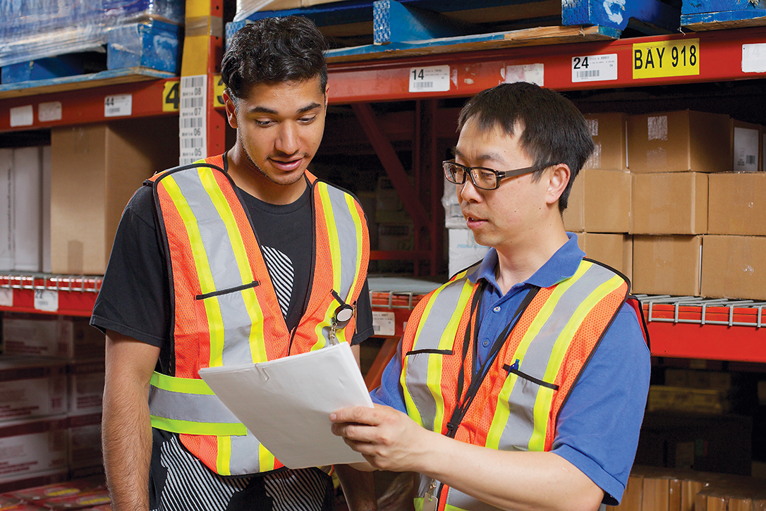 Photo of two males wearing PPE in a warehouse discussing information on a paper document.