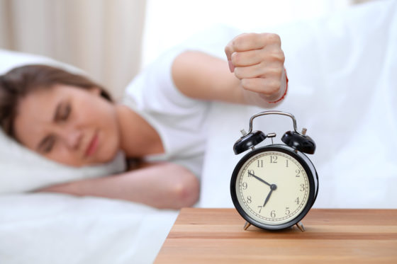 Photo of woman in bed reaching fist toward alarm clock