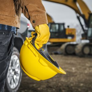 Photo of person holding a yellow hard hat with an excavator in the background.