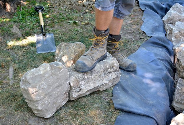 Photo of feet and work boots of a construction worker, with some decorative stone and equipment, that they will use for a landscaping project.